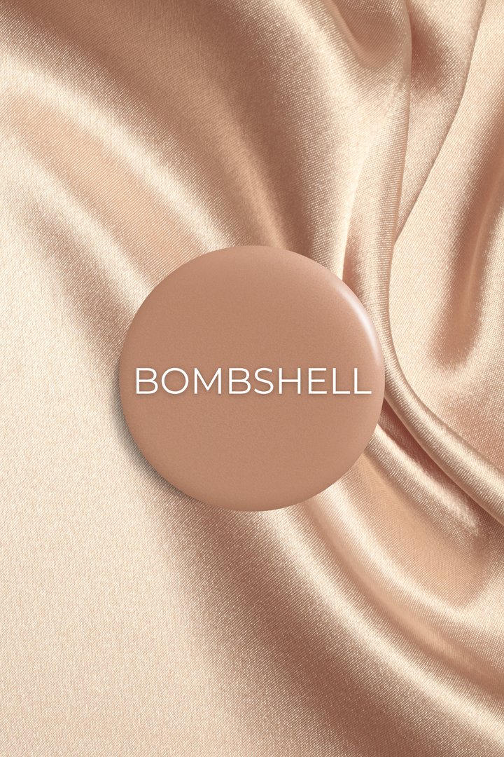 A circular "BOMBSHELL" label rests on luxurious, rippling champagne-colored satin fabric.